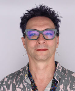 Head shot of Jonathan Gilligan: A White nonbinary person with dark hair, cat-eye glasses, and red lipstick, wearing a pink and gtray patterned shirt and a beaded necklace.